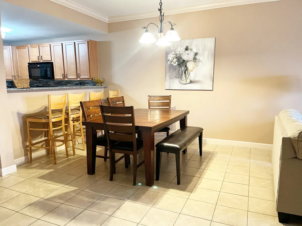 The image centers on a welcoming dining table flanked by bar stools and a sofa, with a decorative painting adding a splash of color. The setup invites shared meals and conversation, highlighting the seamless transition between the dining and living areas.