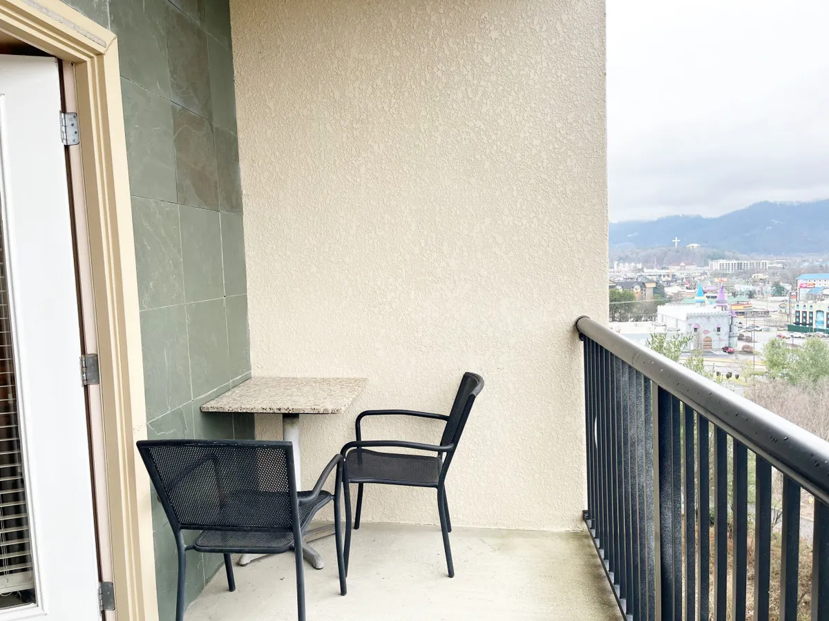 An exterior shot from a balcony or patio reveals a granite table with black chairs, overlooking a sweeping view of the mountains and town below. The serene setting encourages guests to enjoy the scenery immediately after arrival, highlighting the sixth-floor vantage point.