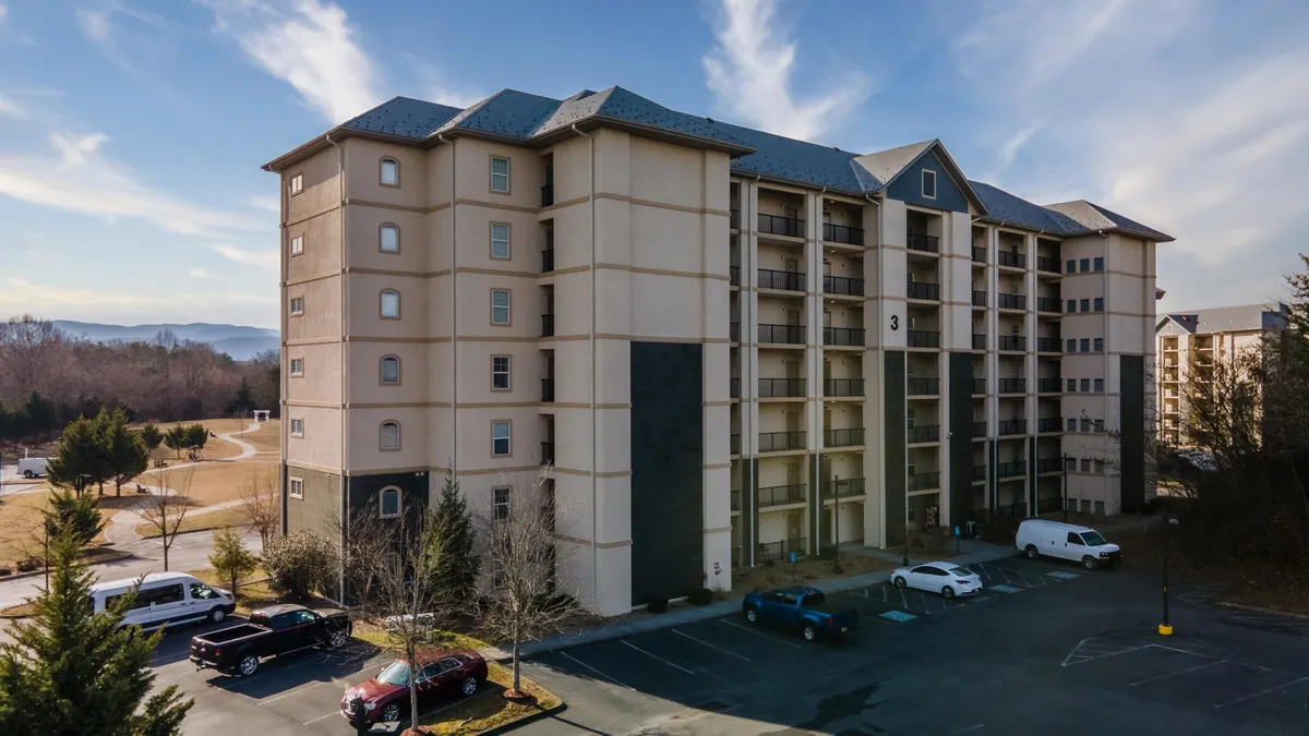 This exterior shot highlights the condo building with its parking lot and lush greenery, suggesting ample outdoor space for gatherings and activities. The welcoming atmosphere is enhanced by the surrounding landscape, hinting at the green spaces perfect for picnics or group games.