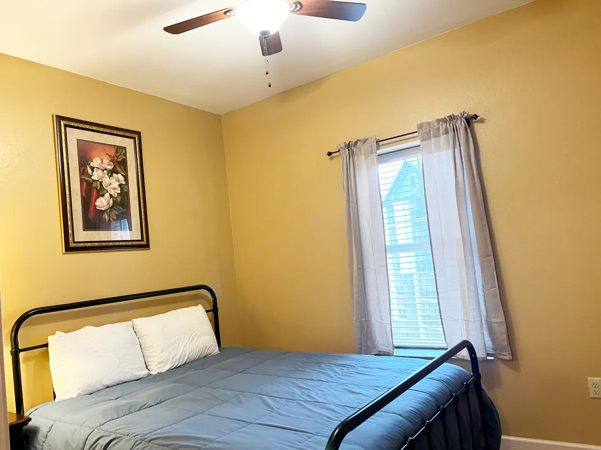 A guest bedroom with a bed dressed in a blue comforter, floral artwork above the headboard, and a window with sheer curtains that filters gentle daylight. The scene is calm and inviting, perfect for relaxation.