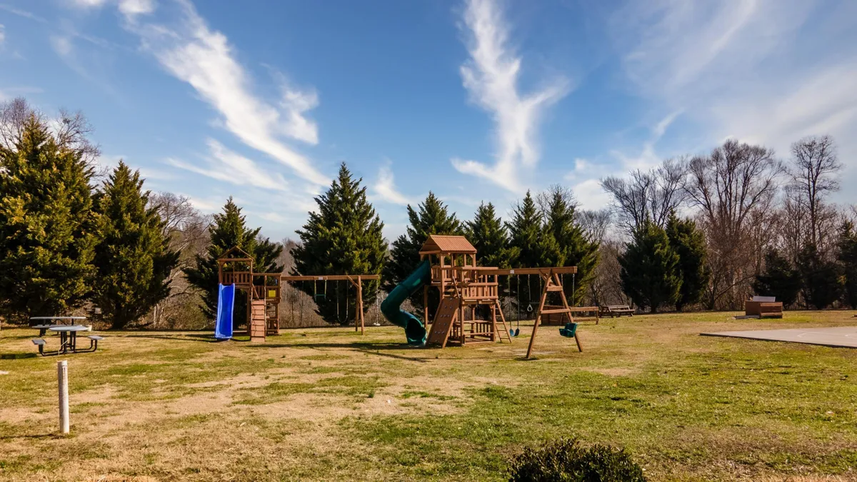 A cheerful outdoor playground scene featuring swings, a slide, green grass, and picnic tables under the open sky. Children play and families gather, creating a vibrant, welcoming atmosphere.