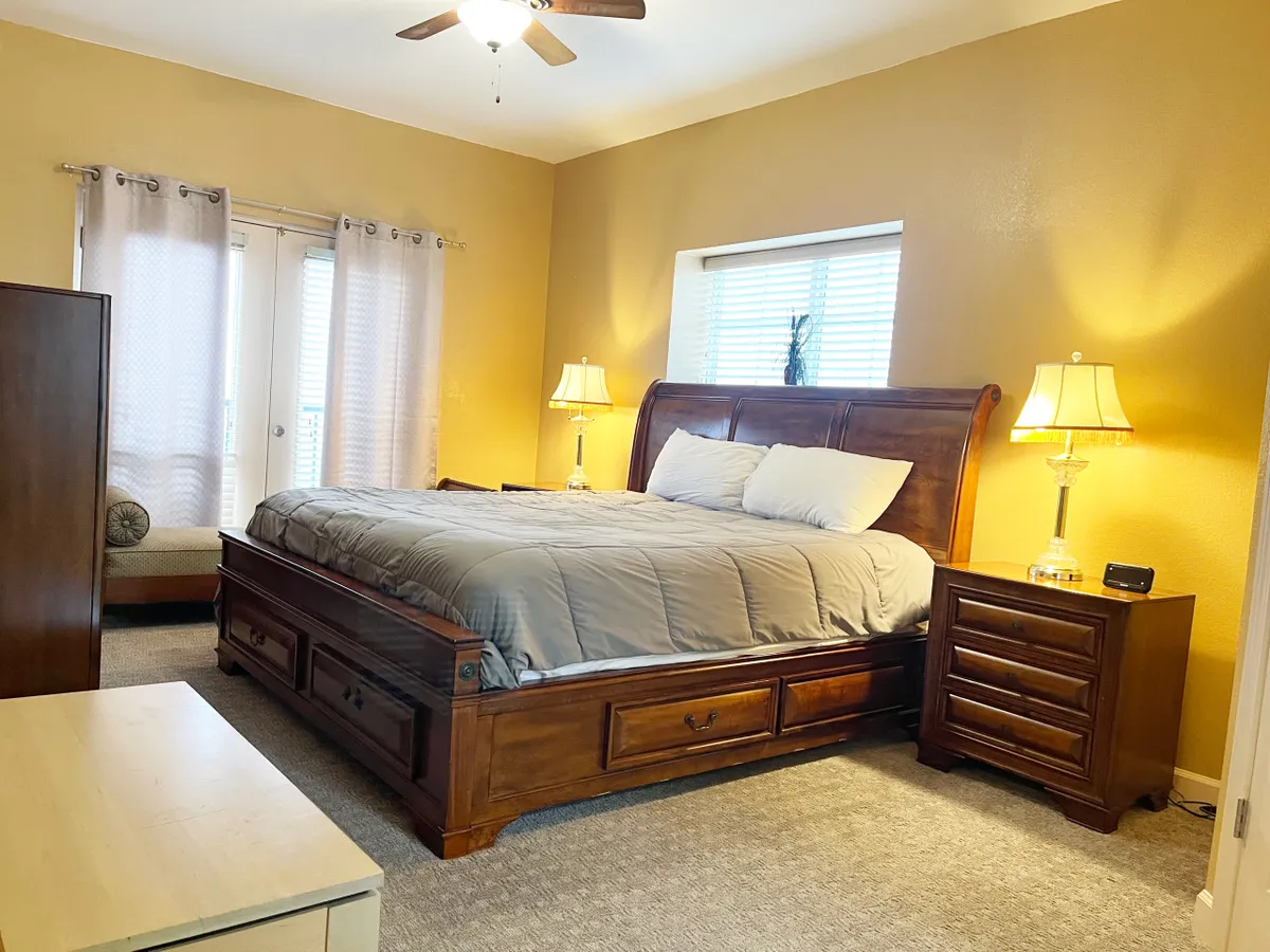 A spacious master bedroom with a king-sized bed, two nightstands with lamps, and a window with curtains letting in natural light. The room exudes warmth and tranquility, ideal for restful nights or peaceful mornings.