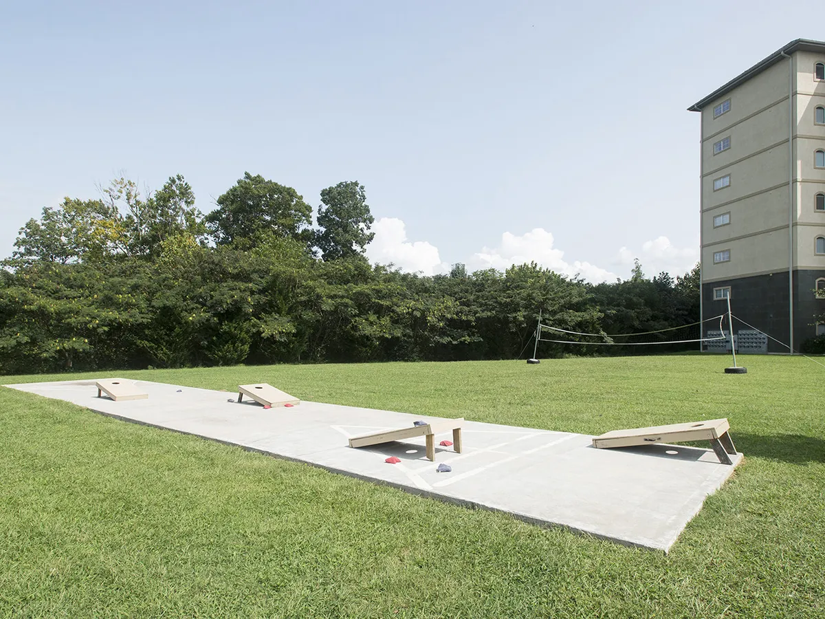 The image showcases the exterior grounds of the condo property, featuring a spacious grassy area set up for bean bag toss and volleyball, bordered by lush trees, with the condo building visible in the background. The scene is inviting and set up for outdoor activities, suggesting opportunities for socializing, playing games, and enjoying the fresh air in a lively, communal atmosphere.